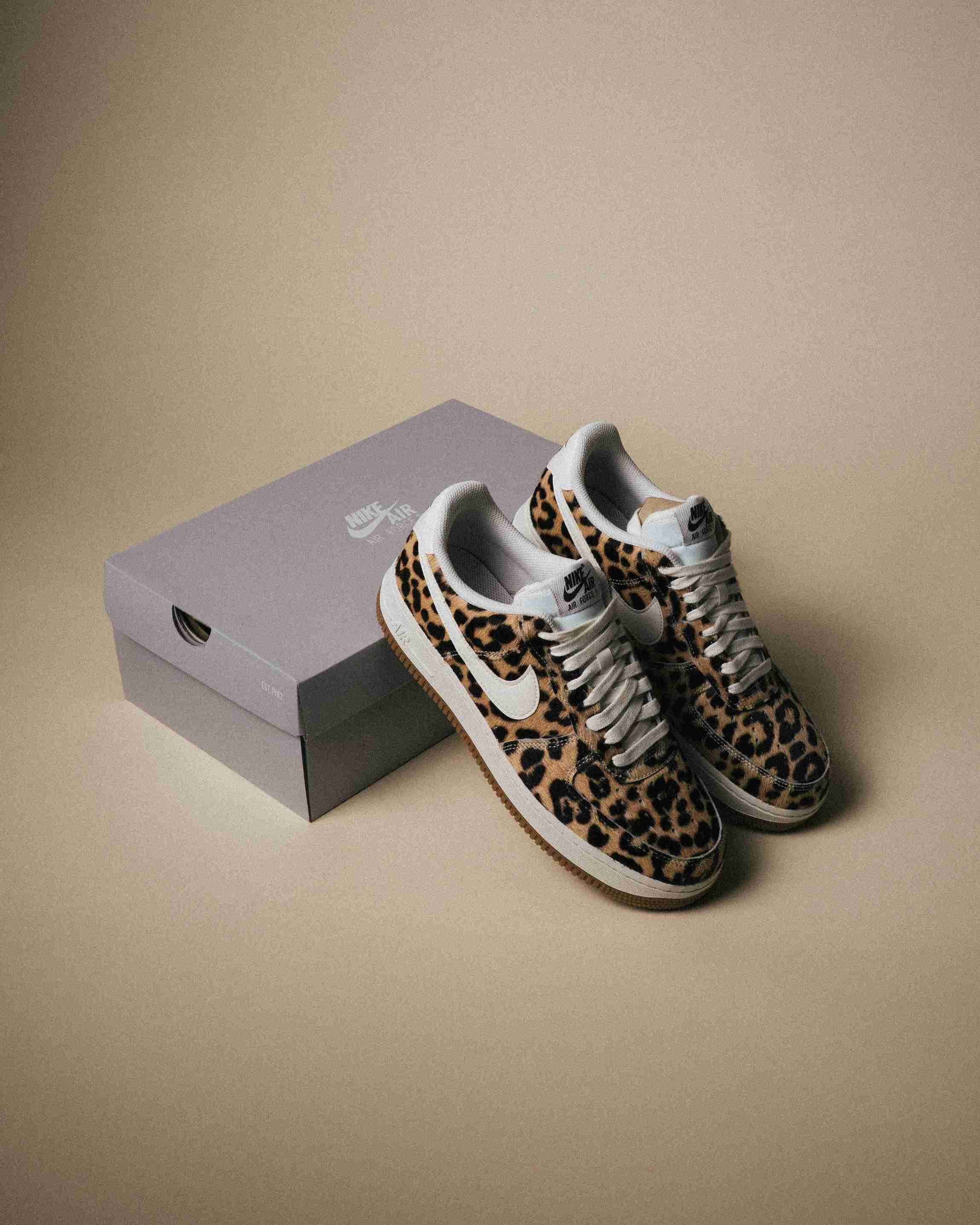 Paire de sneakers Nike Air Force 1 à motif léopard, placée à côté d’une boîte Nike grise sur un fond beige, vue artistique avec lumière douce.